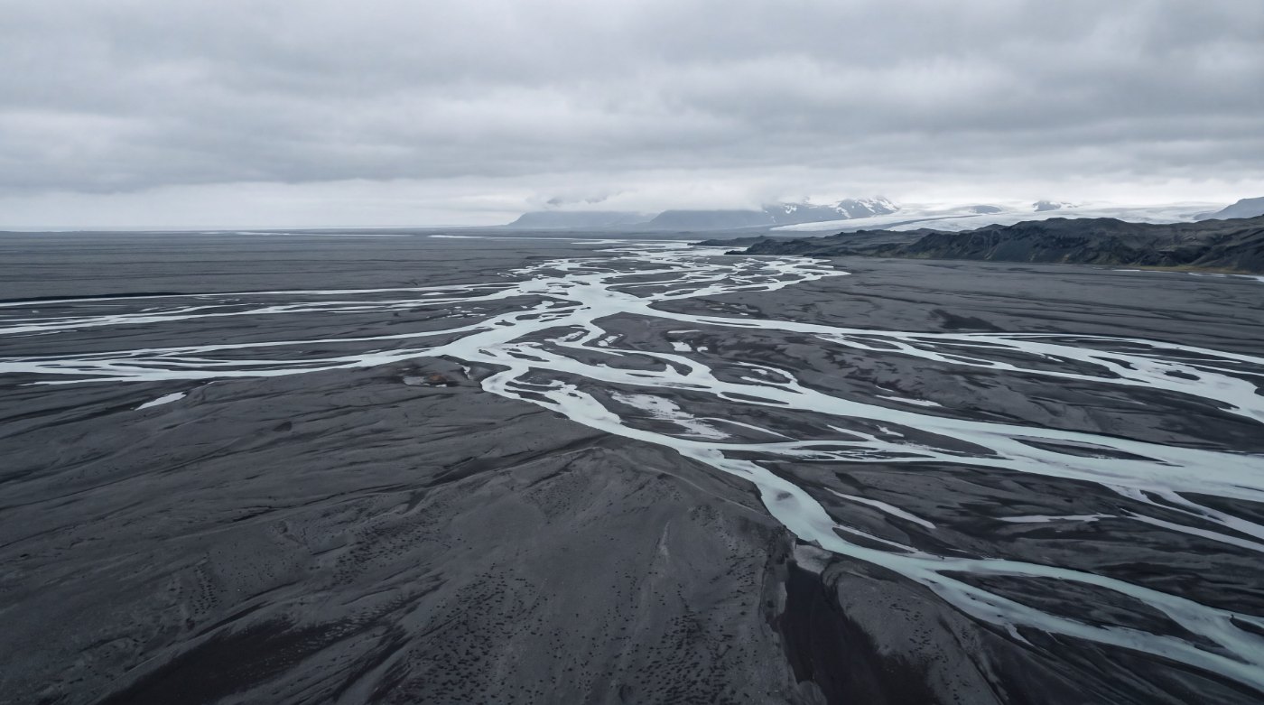 Aerial view of a braided glacial river, many channels converging across a dark plain